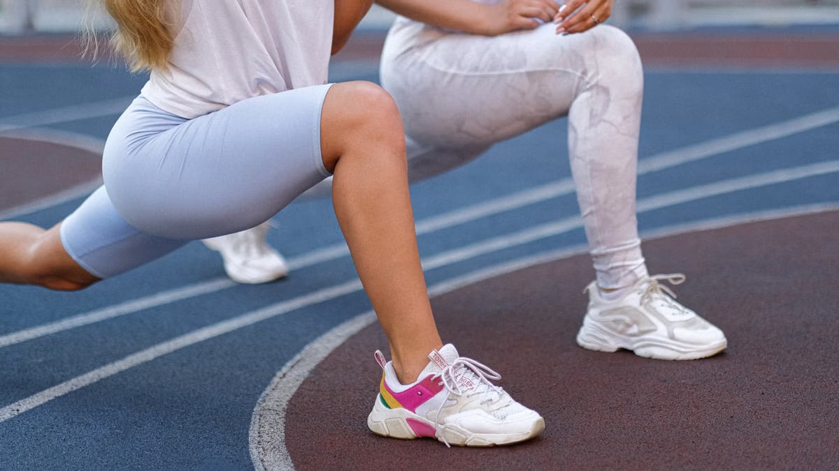 Side view of unrecognizable female athletes in sportswear and sneakers stretching legs doing exercises on stadium